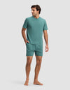 A man stands barefoot against a plain background, wearing the Cozy Earth Men's First Layer Pajama Short Sleeve Tee & Short Set in teal. Facing slightly sideways with one hand in his shorts pocket, he looks relaxed and casual.|Color:Sea Green