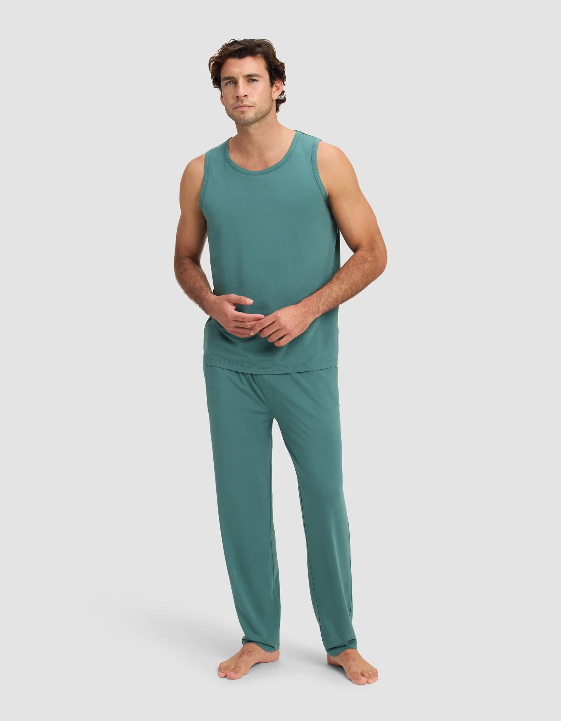A man stands barefoot against a plain background, wearing the Cozy Earth Men's First Layer Pajama Tank & Pant Set in teal. He faces forward with a relaxed expression, one hand touching the other.