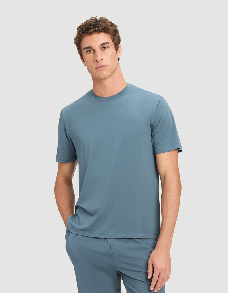 A young man with short curly hair wears the Cozy Earth Men's First Layer Pajama Short Sleeve Tee and matching shorts, standing in front of a plain light gray background with one hand in his pocket.