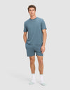 A man stands against a plain background wearing Cozy Earth's Men's First Layer Pajama Short Sleeve Tee in blue, matching blue shorts, and white socks with light blue toes and heels. He has one hand in his pocket and a neutral expression.|Color:Dawn