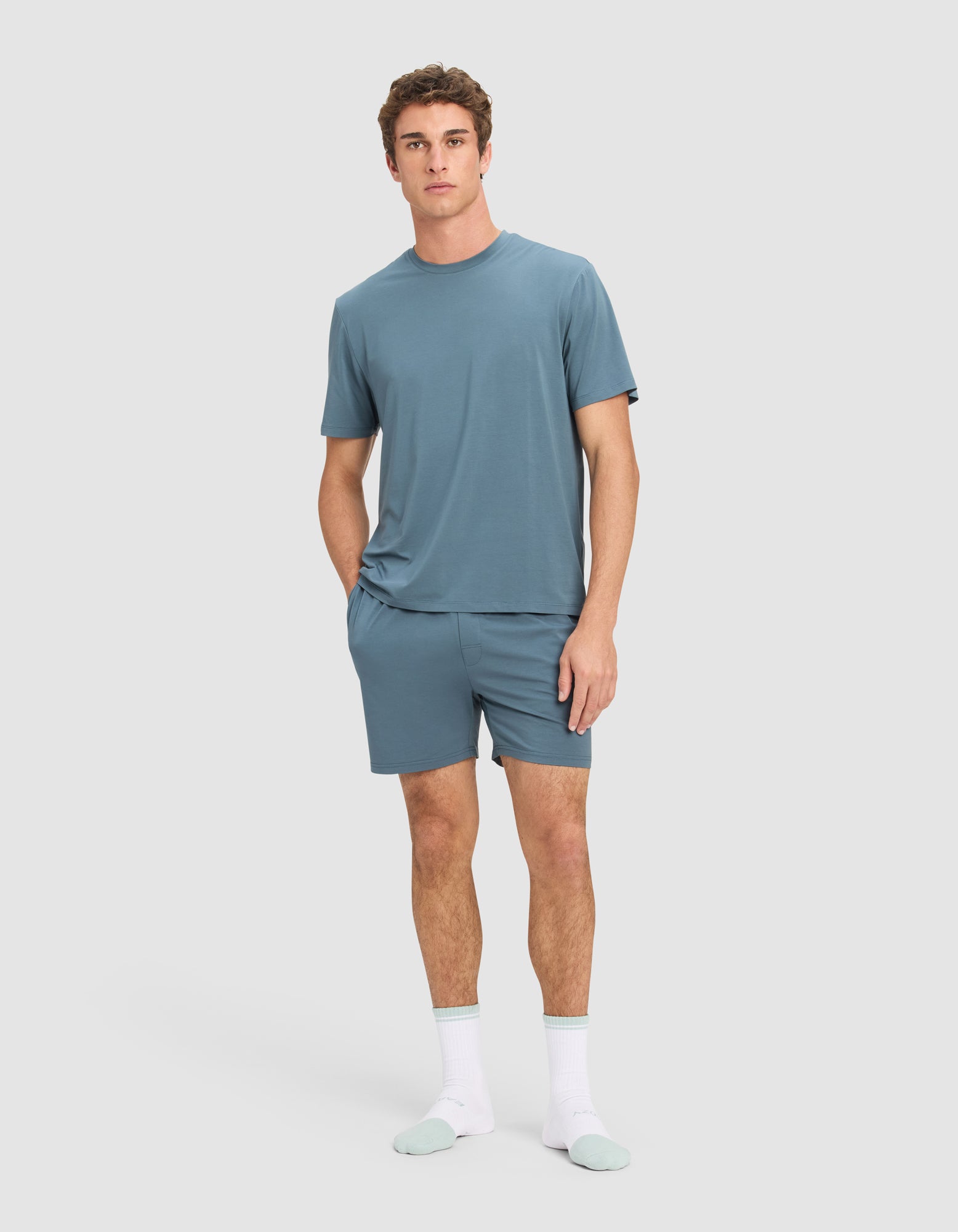 A man stands against a plain background wearing Cozy Earth's Men's First Layer Pajama Short Sleeve Tee in blue, matching blue shorts, and white socks with light blue toes and heels. He has one hand in his pocket and a neutral expression.|Color:Dawn