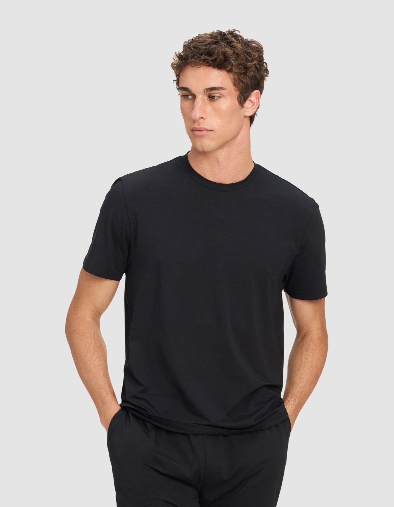 A man wears the Cozy Earth Men's First Layer Pajama Short Sleeve Tee in black, paired with black shorts, standing with hands in his pockets and looking to the side against a light gray background.