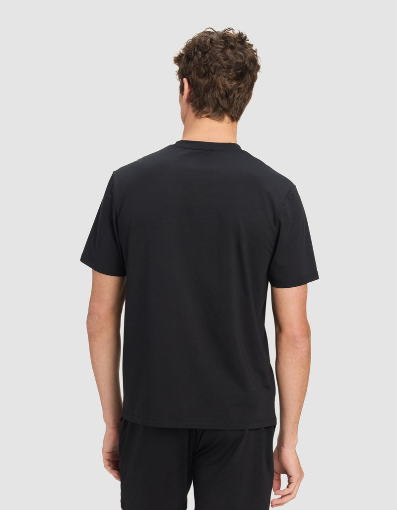 A man with curly brown hair stands against a light gray background, facing away from the camera, wearing a black Men's First Layer Pajama Short Sleeve Tee by Cozy Earth and black shorts.