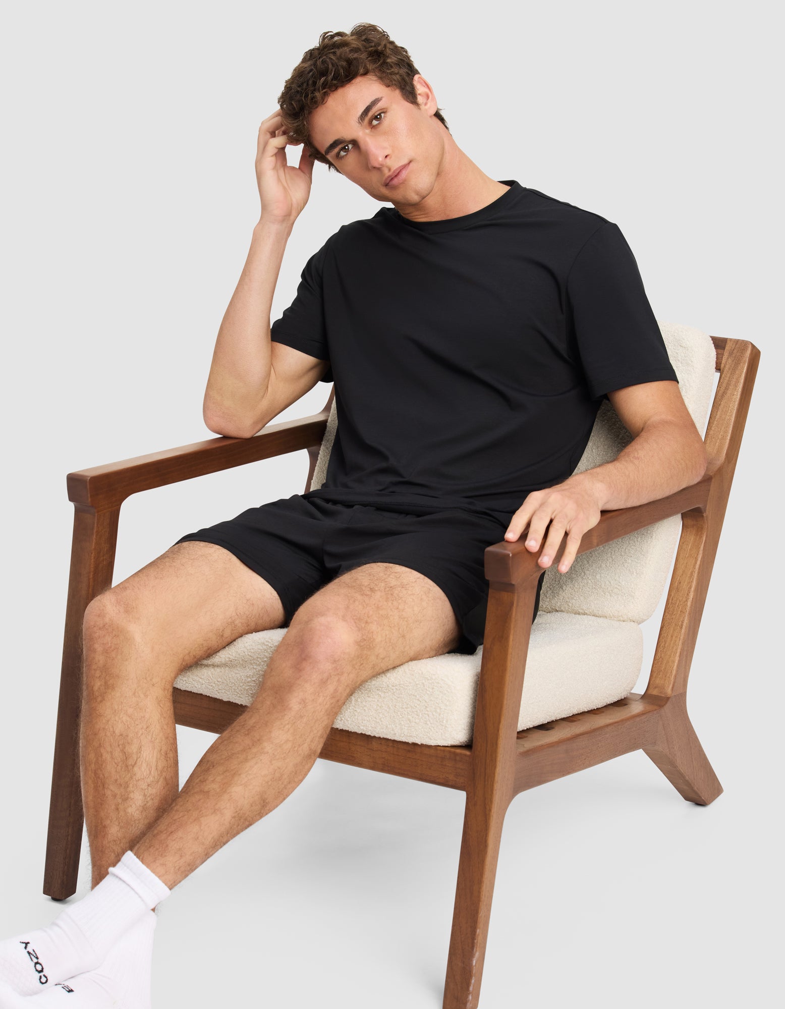 A young man wears the Cozy Earth Men's First Layer Pajama Short Sleeve Tee and shorts as he relaxes in a modern wooden chair with cream cushions, resting one hand on his head and the other on the armrest, paired with white socks.