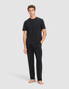 A man stands barefoot against a plain background, wearing the Cozy Earth Men's First Layer Pajama Short Sleeve Tee & Pant Set in black with his left hand in his pocket.|Color:Jet Black