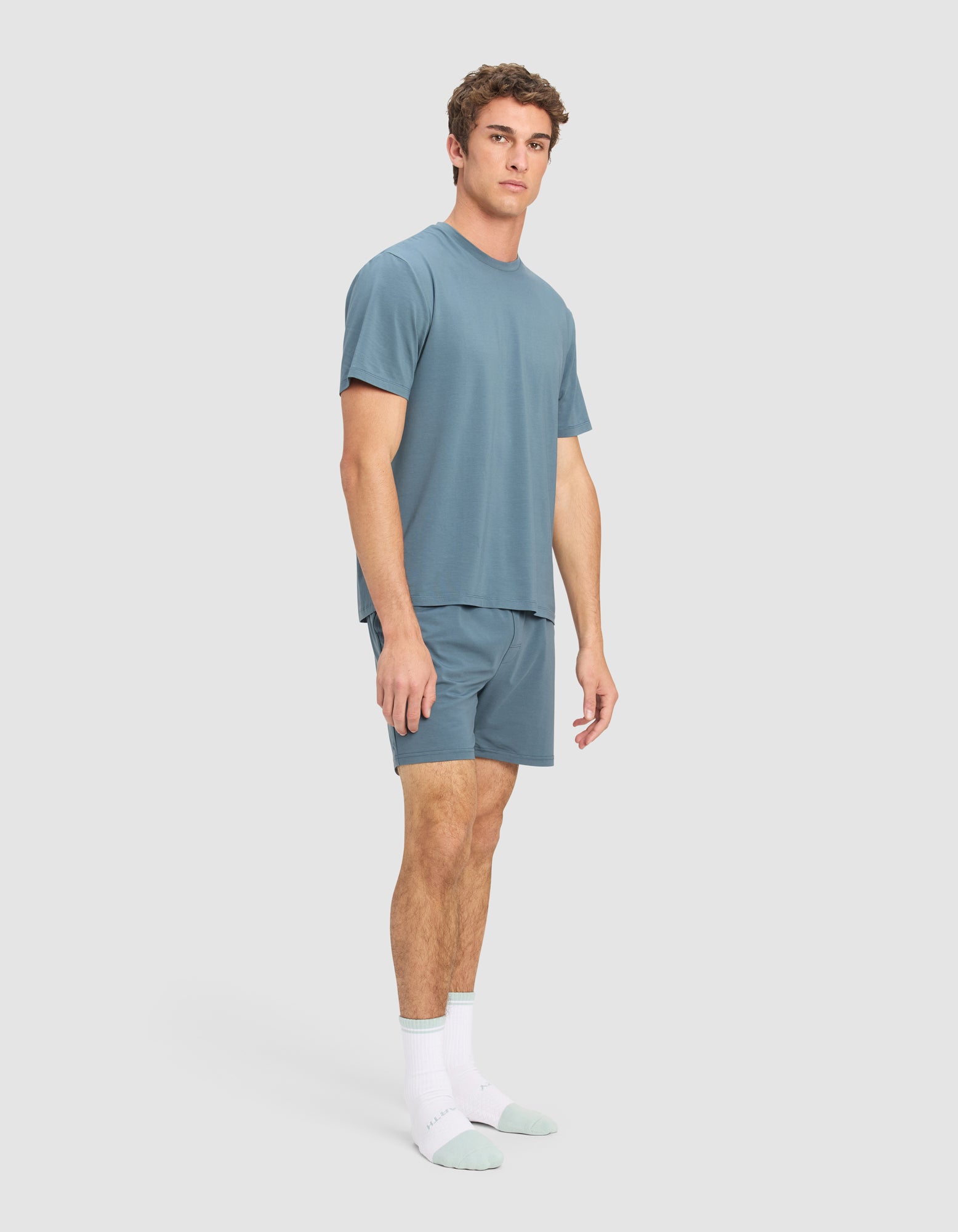 A man stands in a relaxed pose wearing the Cozy Earth Men's First Layer Pajama Short Sleeve Tee & Short Set and white socks with light green toes and heels, against a plain light gray background.