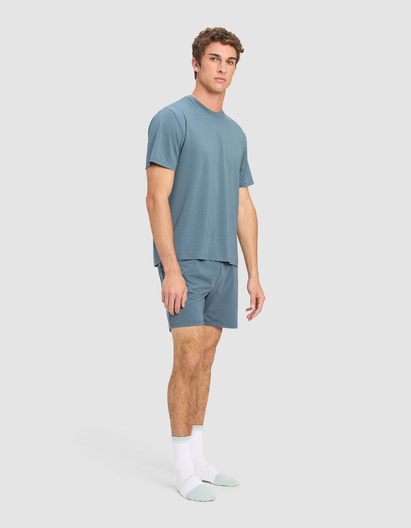 A man stands in a relaxed pose wearing the Cozy Earth Men's First Layer Pajama Short Sleeve Tee & Short Set and white socks with light green toes and heels, against a plain light gray background.
