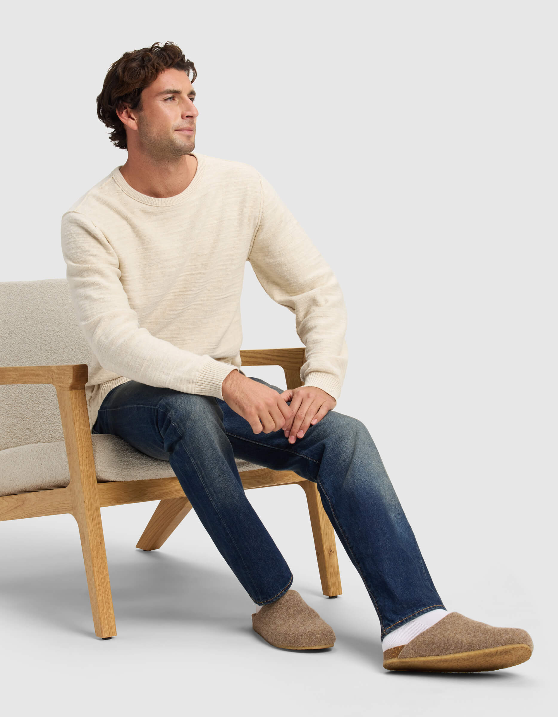 A man in a cream sweater and blue jeans sits on a light wooden chair, wearing Cozy Earth Lakehouse Clogs and gazing to the side with a neutral expression.