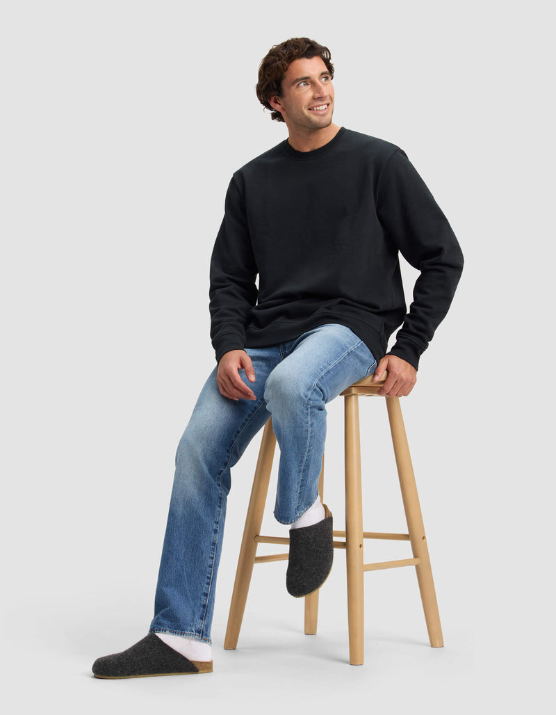 A man with curly brown hair sits on a wooden stool, smiling up, in a black sweatshirt, light blue jeans, white socks, and Cozy Earth Lakehouse Clog slippers. The backdrop is a plain light gray.