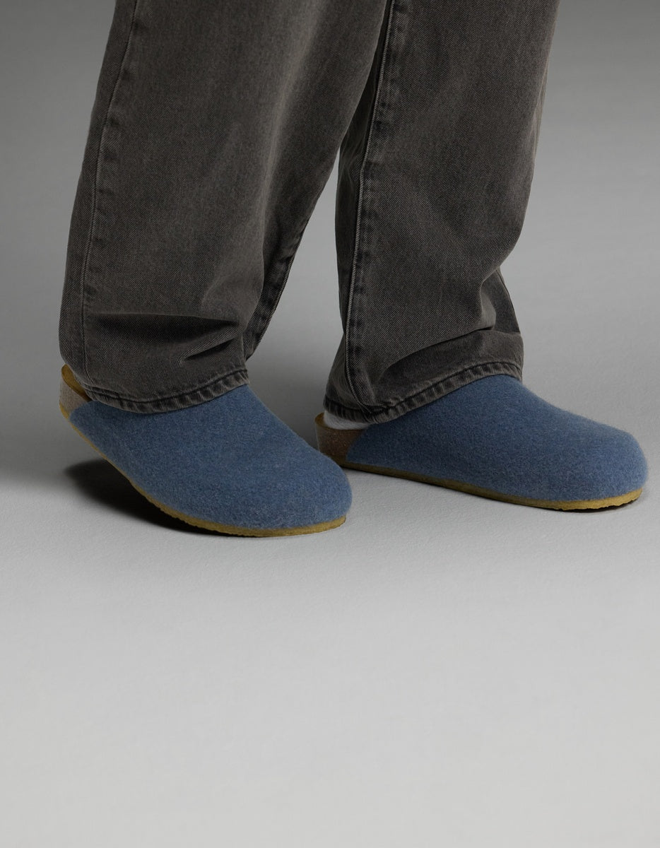 A person wearing dark gray pants and Cozy Earth Lakehouse Clogs in Light Blue stands on a light gray floor, with only their lower legs and feet visible.|Color:Light Blue