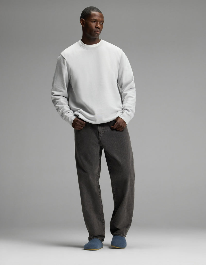 A man stands against a plain gray background, wearing a light gray sweatshirt, loose dark gray jeans, and Cozy Earth’s Lakehouse Clog slippers in blue. His hands are in his pockets and he has a neutral expression.