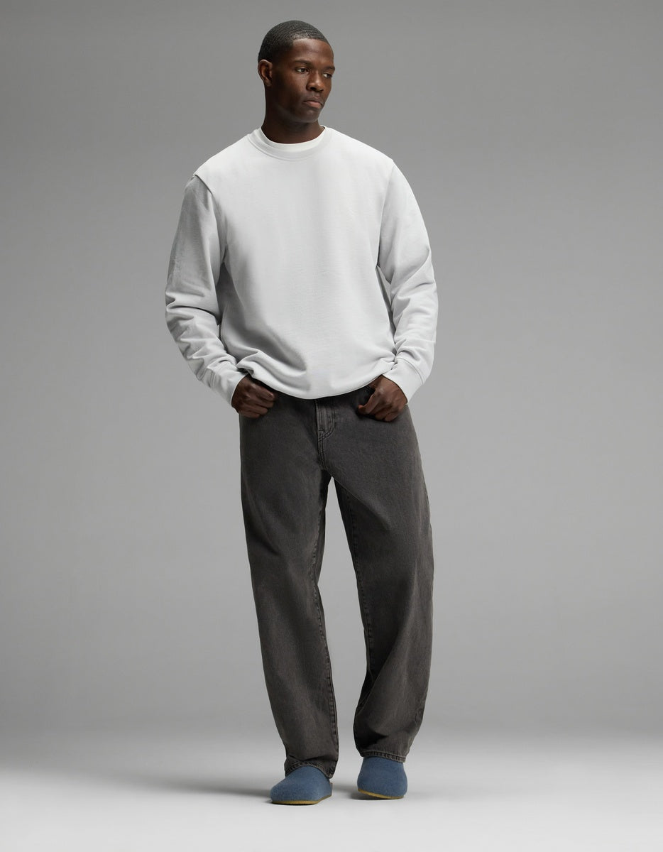 A man stands against a plain gray background, wearing a light gray sweatshirt, loose dark gray jeans, and Cozy Earth’s Lakehouse Clog slippers in blue. His hands are in his pockets and he has a neutral expression.|Color:Light Blue