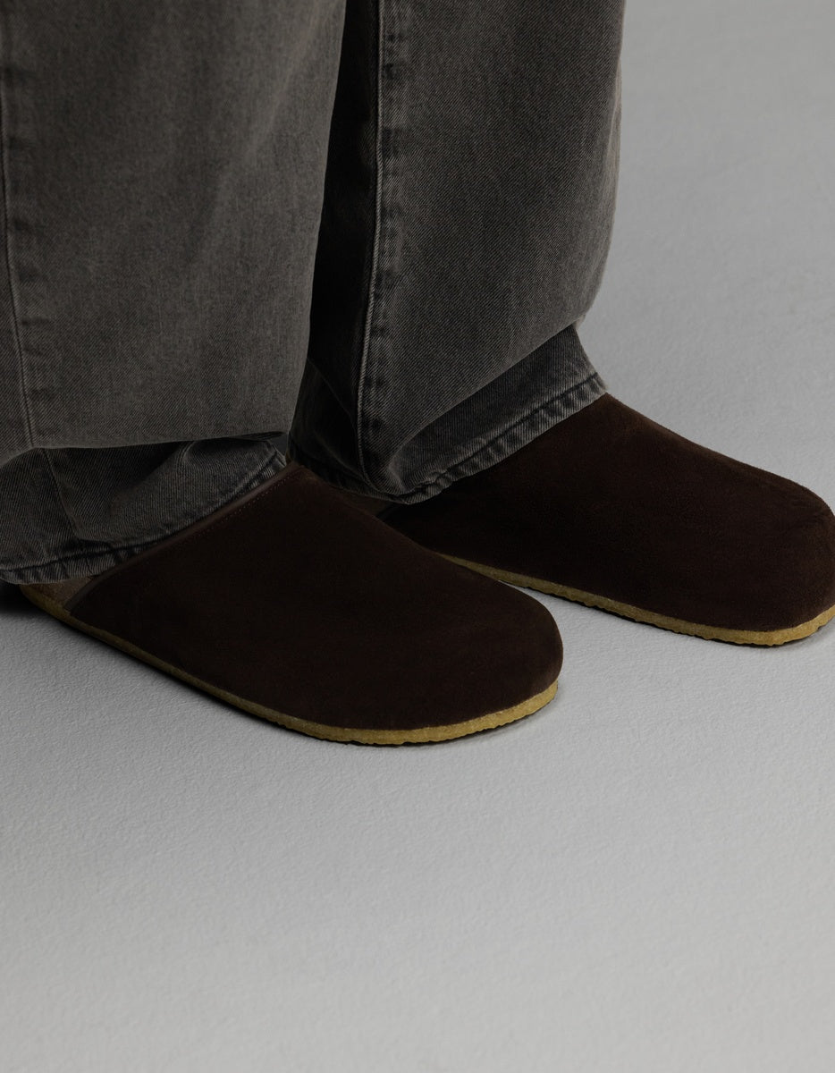 A person wearing the Cozy Earth Lakehouse Clog in Suede—dark brown slippers with light brown soles—paired with gray jeans stands on a light gray floor.