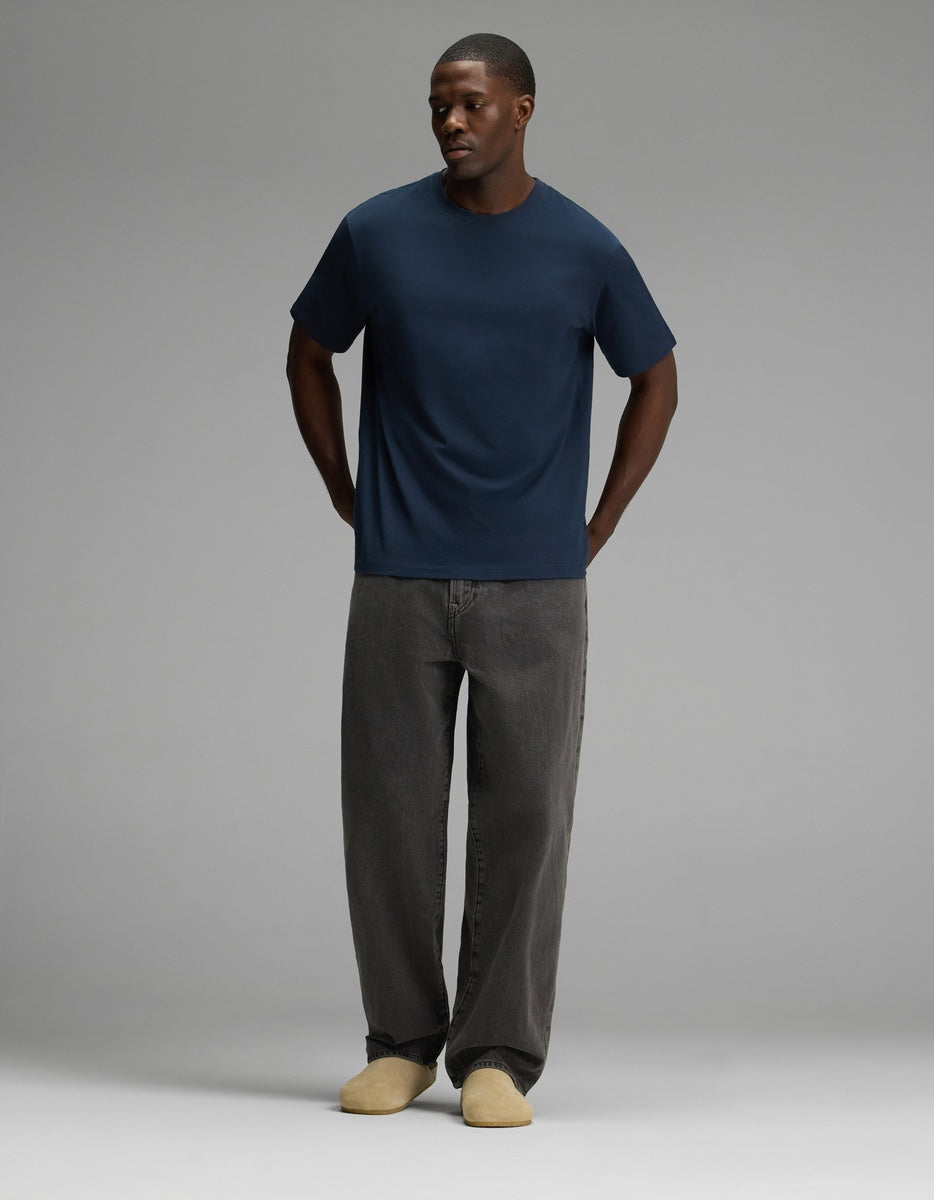 A person stands against a plain gray background, wearing a navy blue t-shirt, loose-fitting dark gray jeans, and the Lakehouse Clog in Suede by Cozy Earth, with their hands in their pockets.