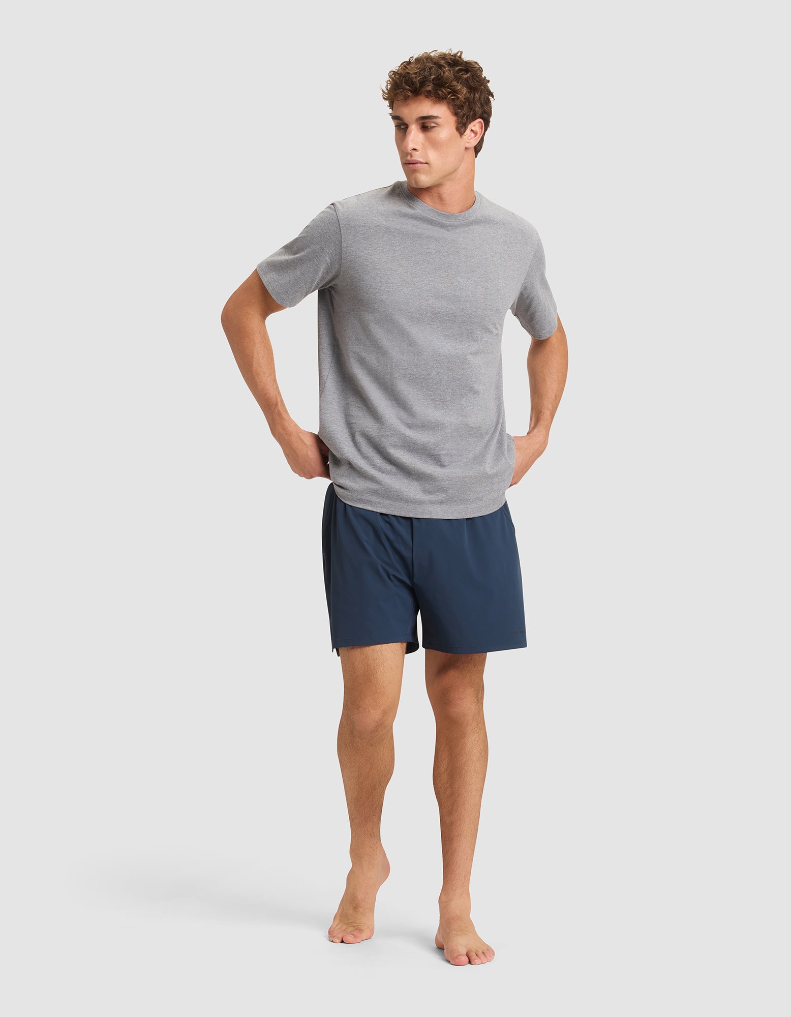 A man stands barefoot against a plain background, wearing a light gray short-sleeve T-shirt and Cozy Earth Men's Day & Night Shorts 5" in dark blue, with his hands resting on his hips. |Color:Eclipse