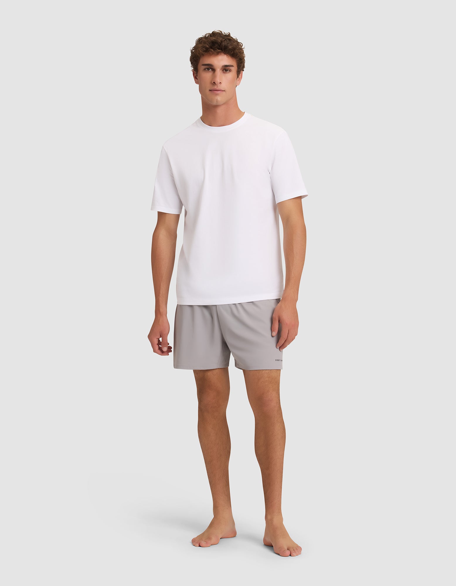 A young man stands barefoot against a light grey background, facing forward with a neutral expression. He is wearing Cozy Earth Men's Day & Night Shorts 5" in light grey and a plain white short-sleeve t-shirt. |Color:Stone