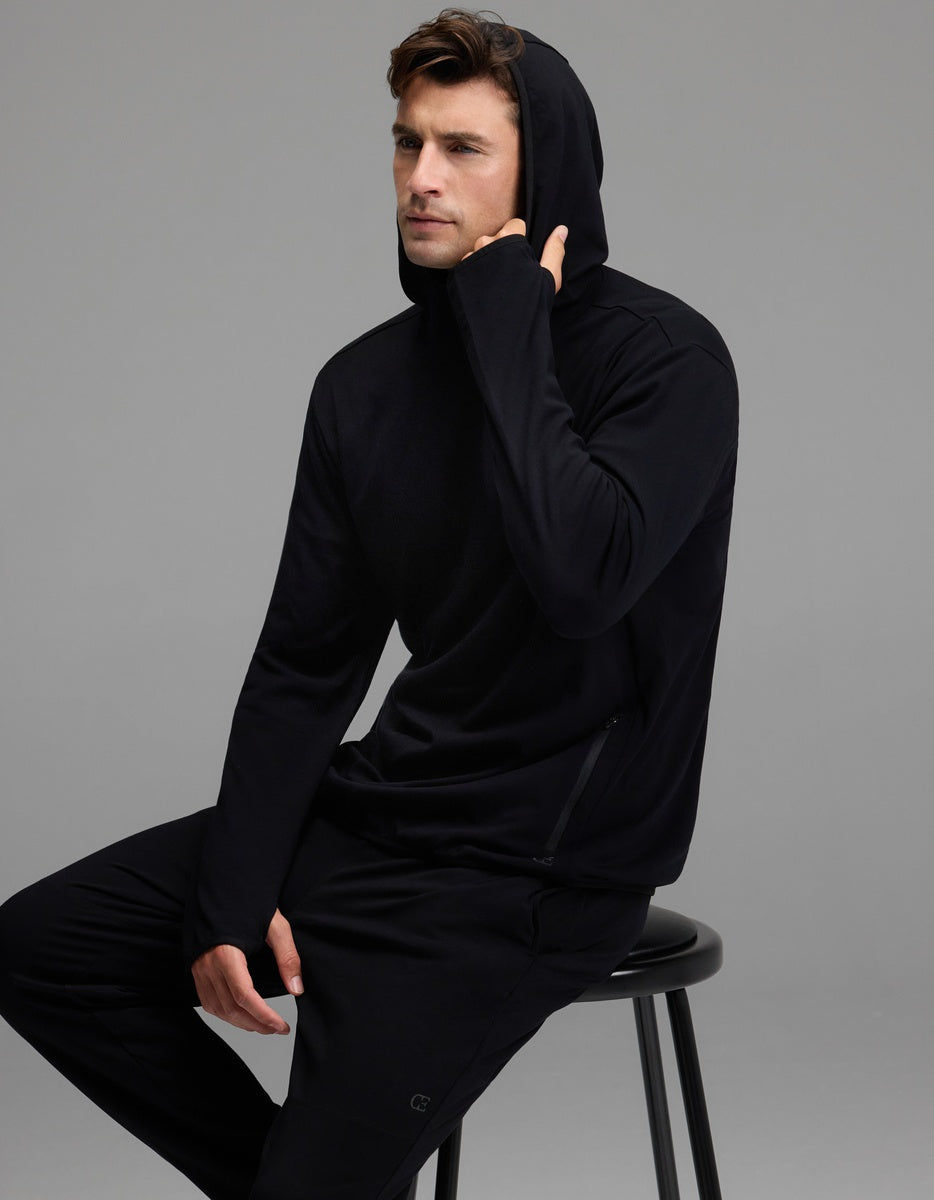 A man wearing the Cozy Earth Men’s Recovery Hoodie and matching pants sits on a black stool against a gray background, with one hand touching his hoodie near his chin.