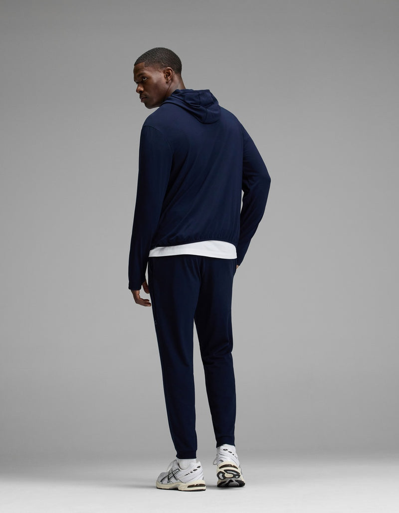 A man wearing Cozy Earth’s Men’s Recovery Jogger in navy, paired with a white T-shirt and white sneakers, stands facing away and glances over his left shoulder against a plain gray background.