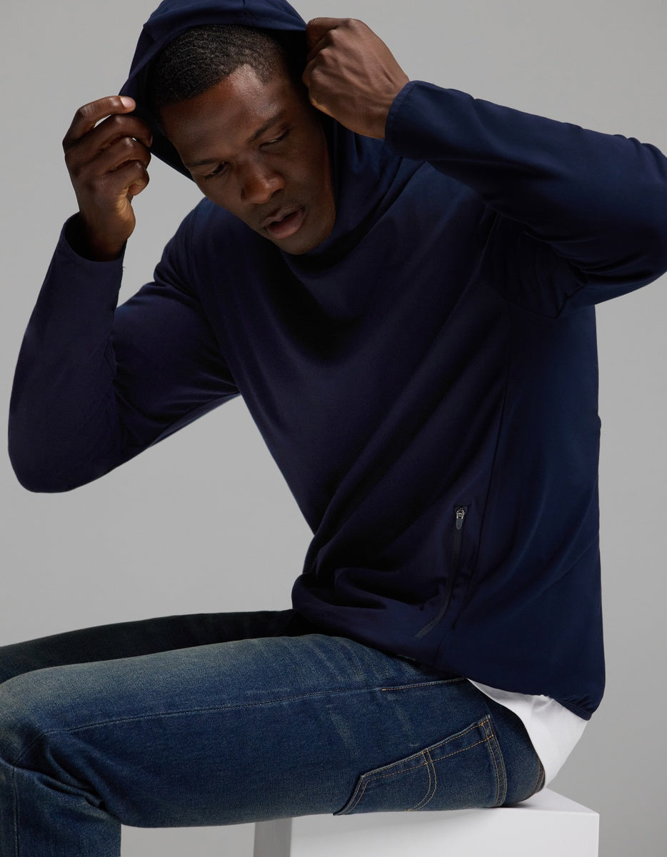 A person wearing the Cozy Earth Men’s Recovery Hoodie and blue jeans sits on a white block, pulling the hood over their head, against a plain gray background.