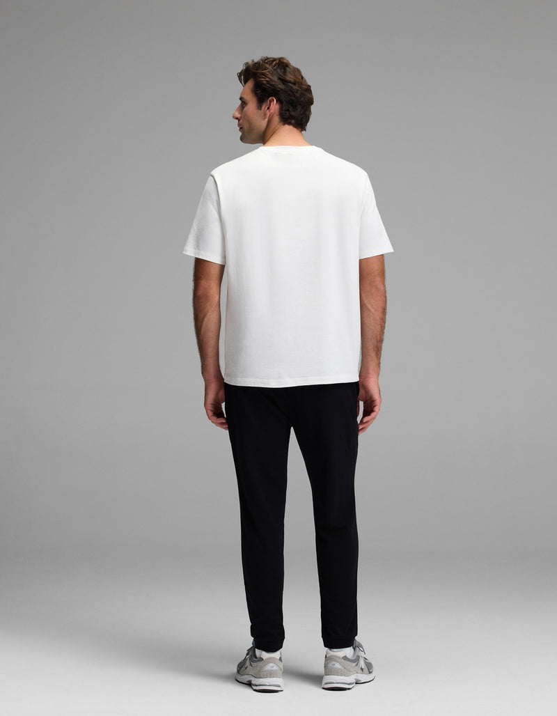 A man stands facing away from the camera, wearing the Cozy Earth Men’s Recovery Jogger with a plain white T-shirt and gray sneakers against a plain gray background.