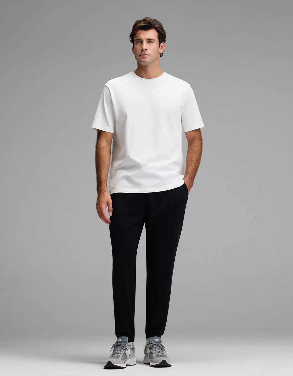 A man stands facing forward against a plain gray background, wearing the Cozy Earth Men’s Recovery Jogger, a plain white t-shirt, and gray sneakers with his hands in his pockets.
