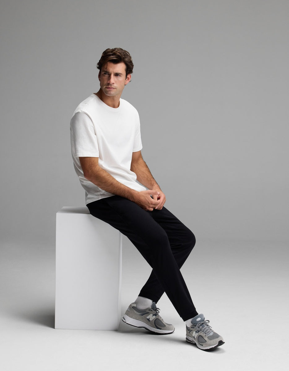 A man in a white t-shirt, Cozy Earth’s Men’s Recovery Jogger in black, and gray sneakers sits on a white block against a plain gray background, looking to the side.