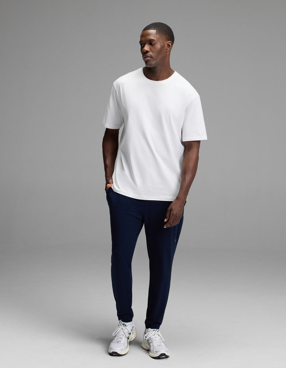 A man stands in a studio wearing a Cozy Earth Men’s Recovery Jogger in dark navy, paired with a plain white t-shirt and white sneakers. He looks slightly to the side with a neutral expression against a plain gray background.