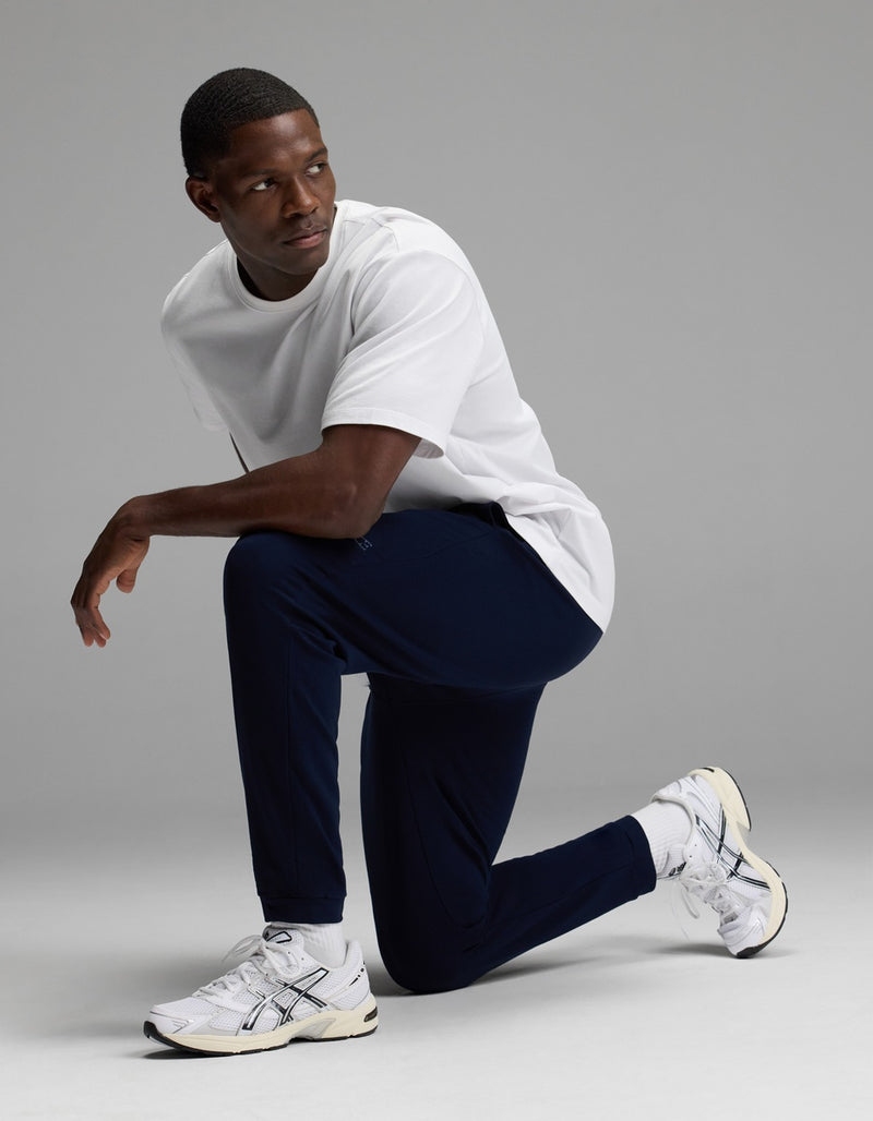 A man wearing a white t-shirt, white sneakers, and Cozy Earth Men’s Recovery Jogger in navy kneels on one knee against a plain gray background, looking to his left.