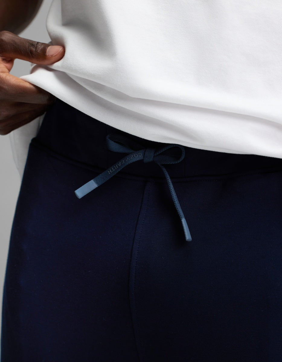 A man wearing Cozy Earth’s Men’s Recovery Jogger in navy blue and a white shirt, adjusting the drawstring waistband with one hand.