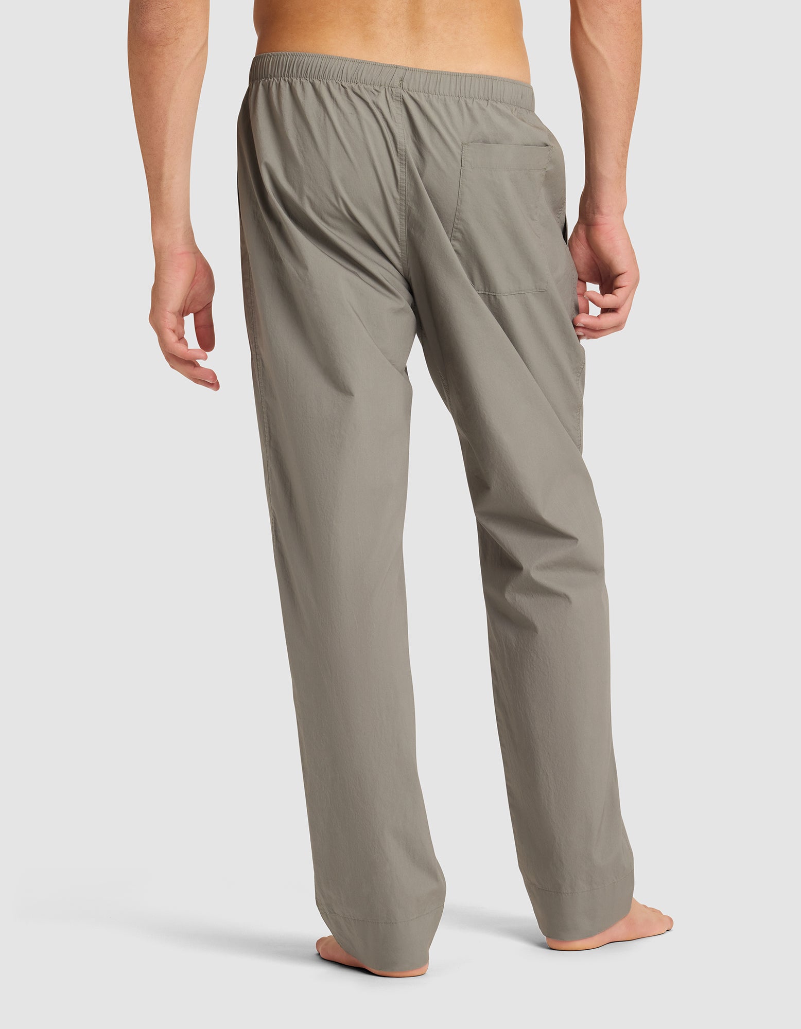 A person stands barefoot, facing away, wearing Cozy Earth Men's Sleep Pant in light gray. The pants are loose-fitting with an elastic waistband and a single right back pocket. Background is plain white. 
