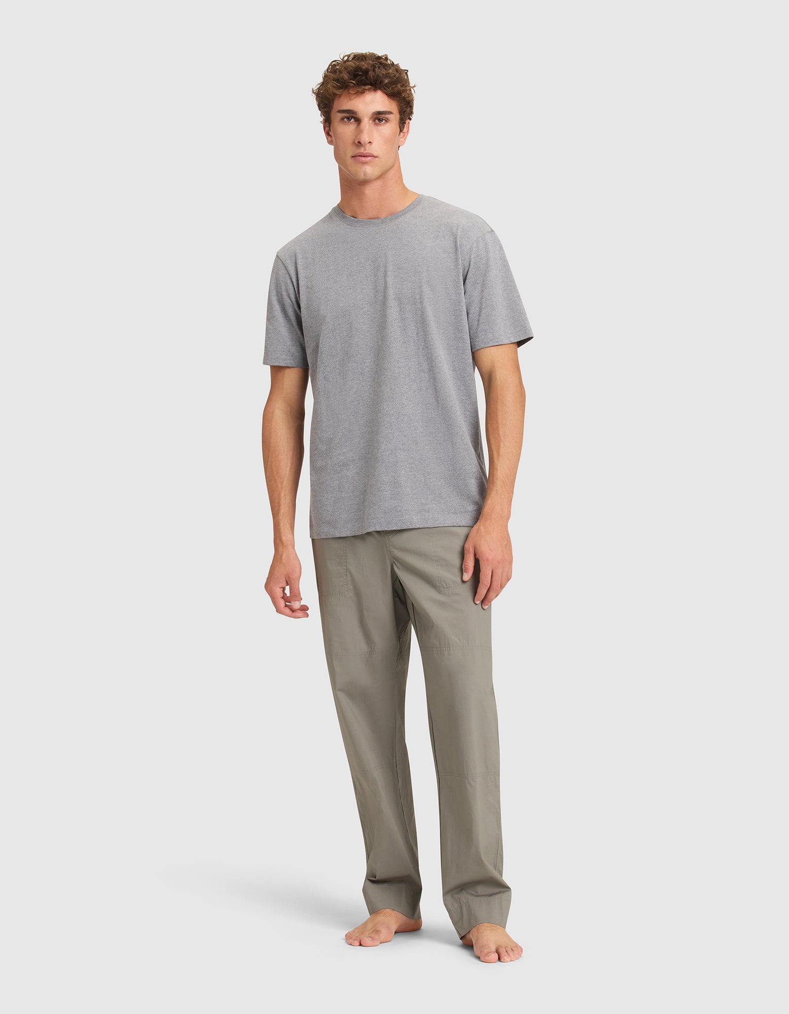 A man with short curly hair stands barefoot against a plain light background, wearing Cozy Earth Men's Sleep Pant and a plain grey T-shirt. 