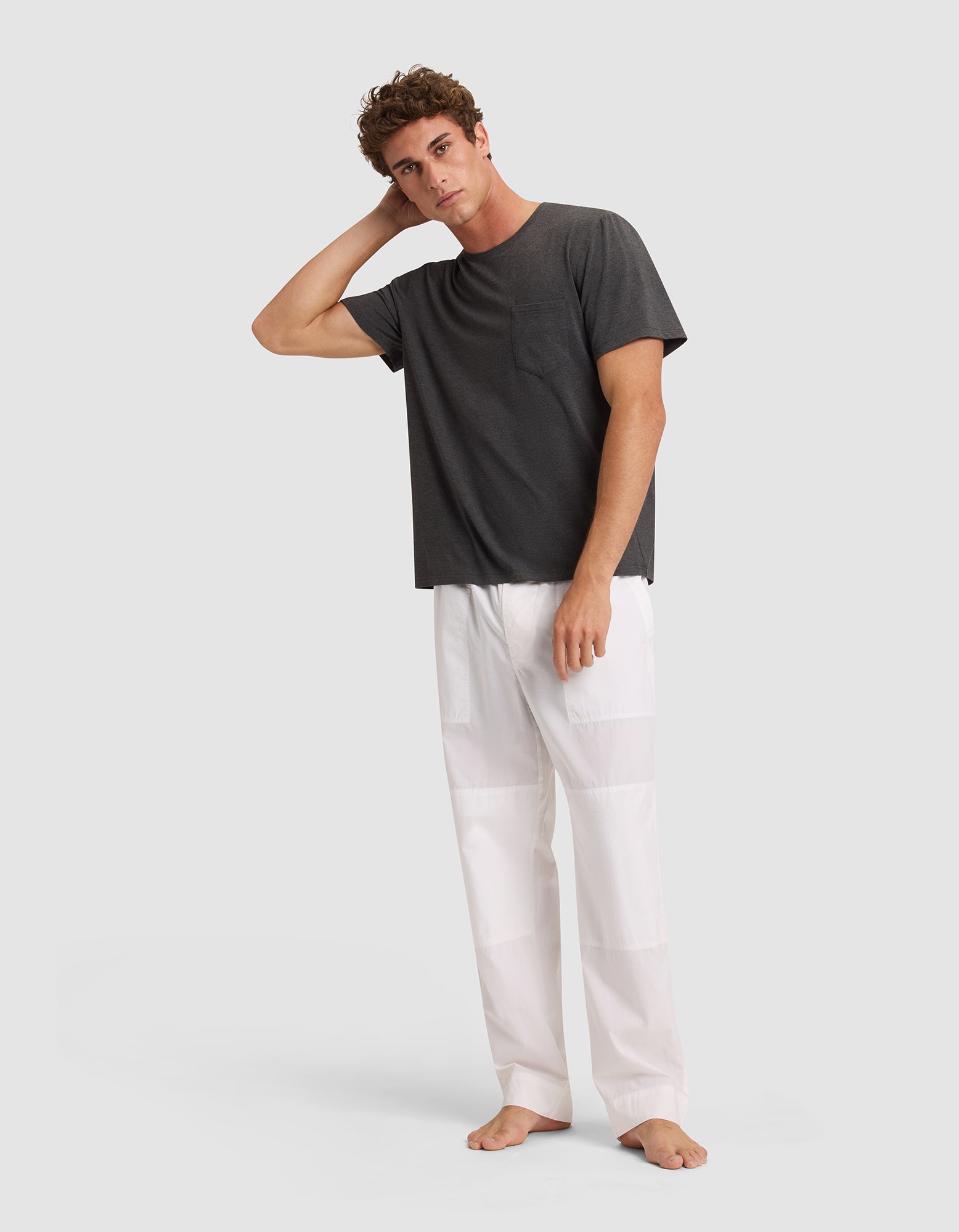 A man stands barefoot against a light background, looking relaxed with one hand on his head, dressed in Cozy Earth Men's Sleep Pant—loose white pants paired with a dark gray t-shirt. |Color:White