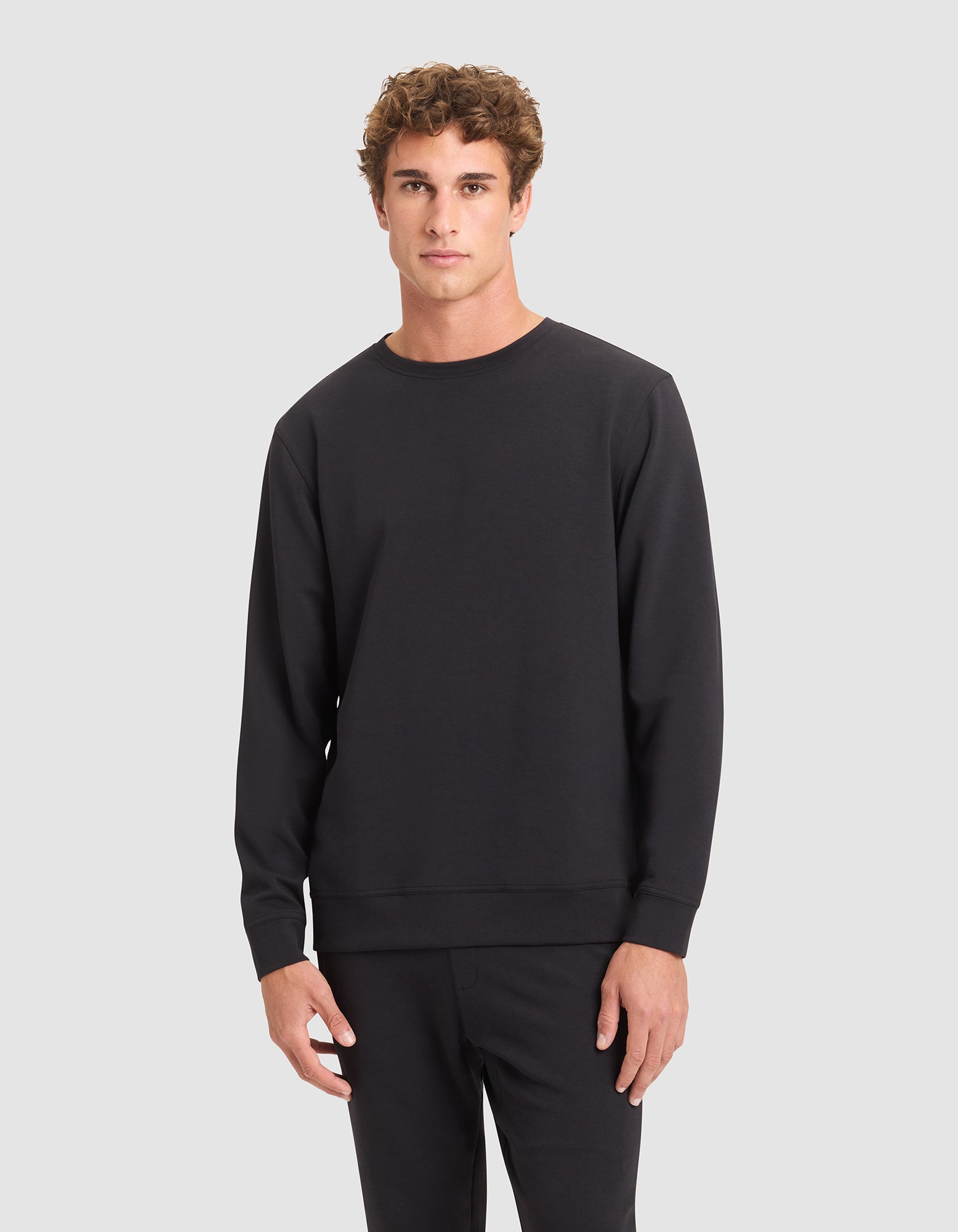 A young man with curly brown hair wears the Cozy Earth Men's StretchTech Crewneck in black with matching black pants, standing against a light gray background. 
