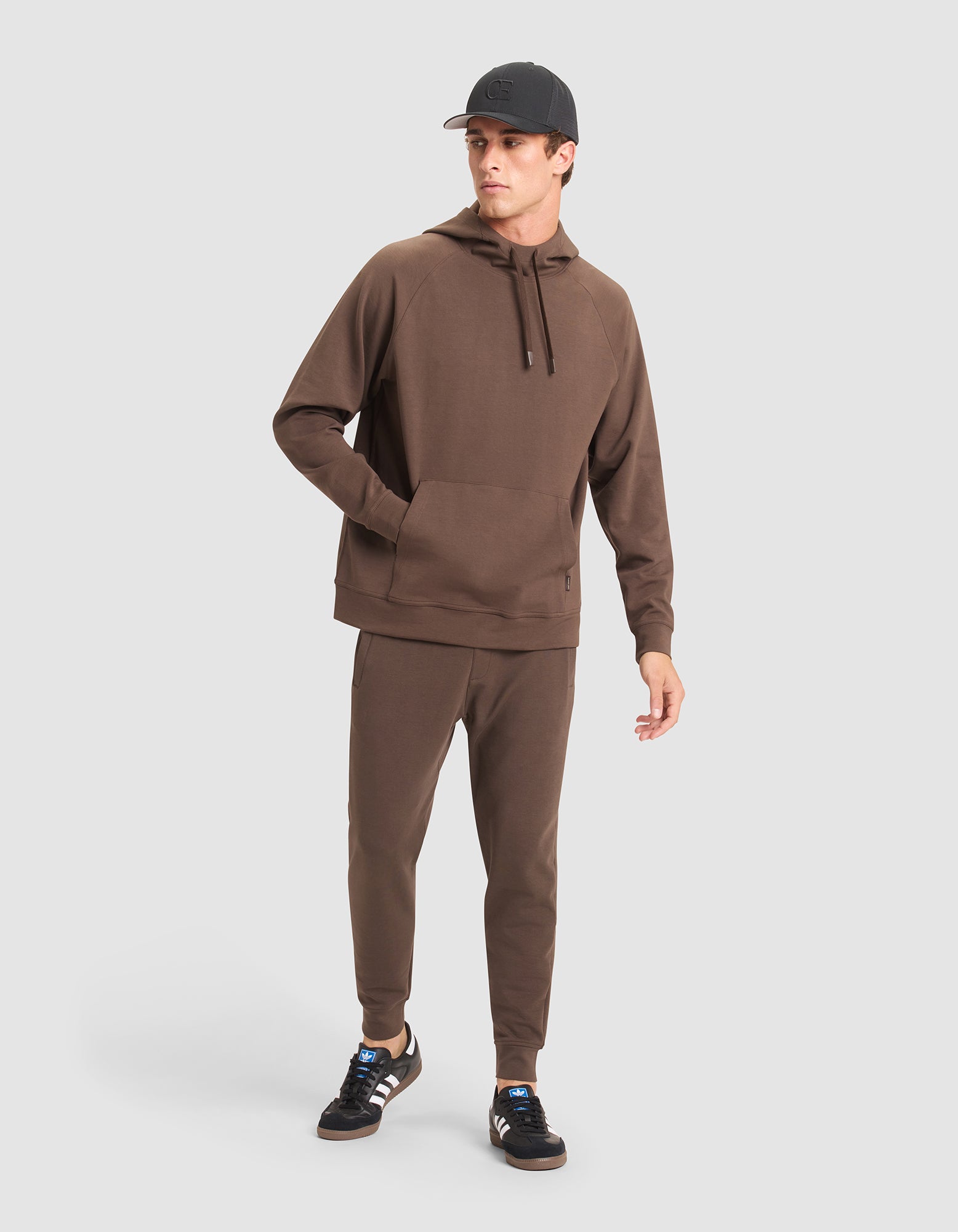 A man wears the Cozy Earth Men's StretchTech Hoodie & Jogger Set with a black cap and black Adidas sneakers featuring white stripes, standing against a plain light gray background. 