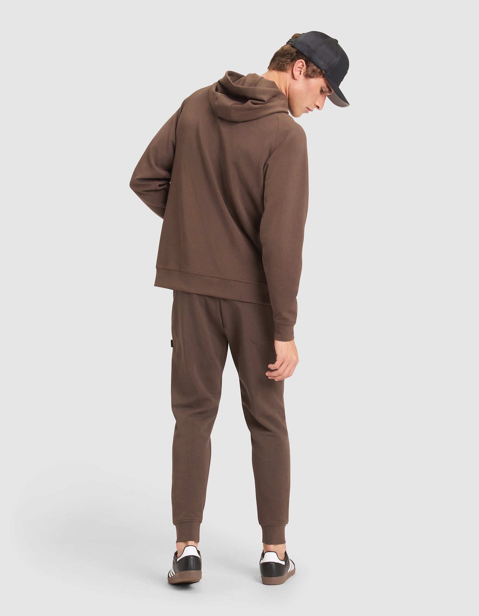 A man models the Cozy Earth Men's StretchTech Hoodie & Jogger Set in brown, paired with a black cap and black-and-white sneakers, standing against a plain light background and glancing over his shoulder.  