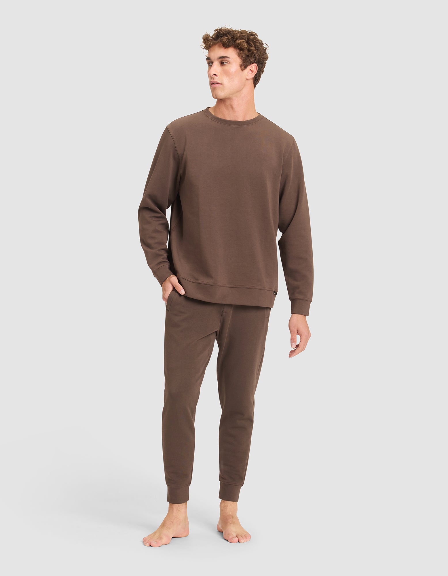 A young man with curly hair stands barefoot against a light gray backdrop, wearing the Cozy Earth Men's StretchTech Crewneck & Jogger Set in brown. 
