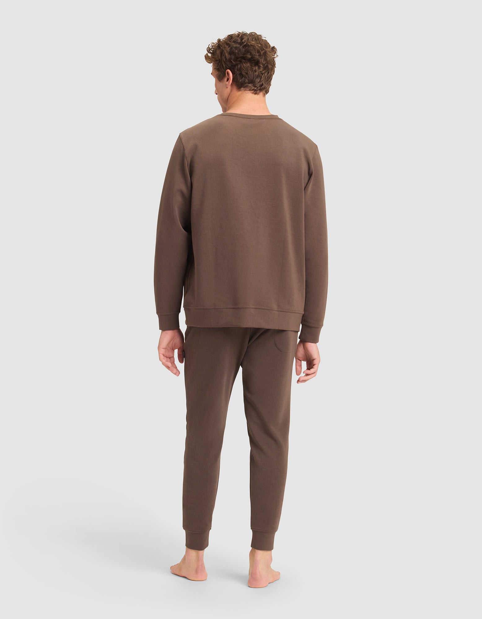 A man stands barefoot facing away, wearing the Cozy Earth Men's StretchTech Crewneck & Jogger Set in brown, set against a plain light gray background. 