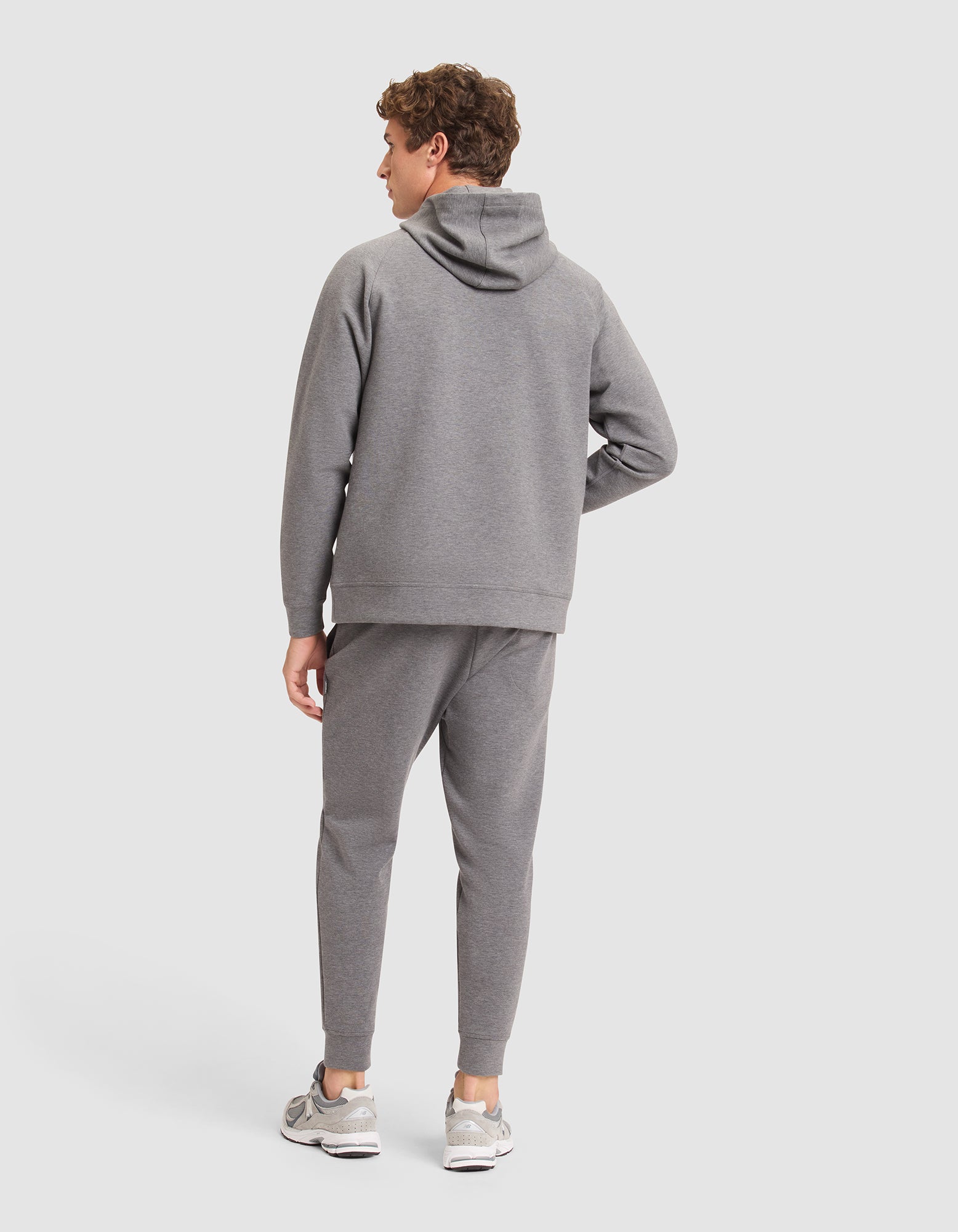A man faces away from the camera, dressed in Cozy Earth's Men's StretchTech Hoodie & Jogger Set in gray, paired with gray sneakers against a plain light background. |Color:Coal Heather
