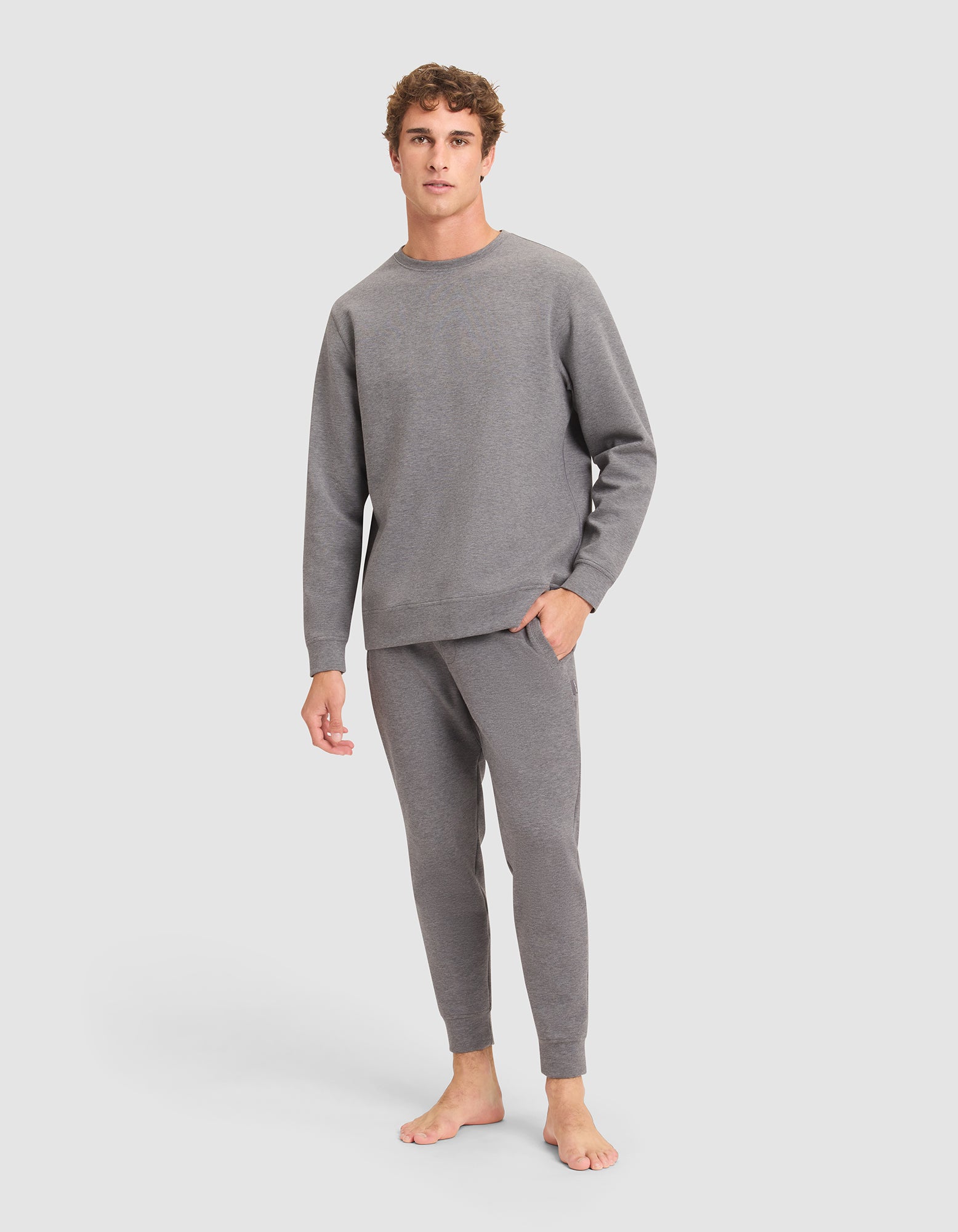 A young man with curly hair stands barefoot against a plain background, wearing the Cozy Earth Men's StretchTech Crewneck & Jogger Set in gray. |Color:Coal Heather