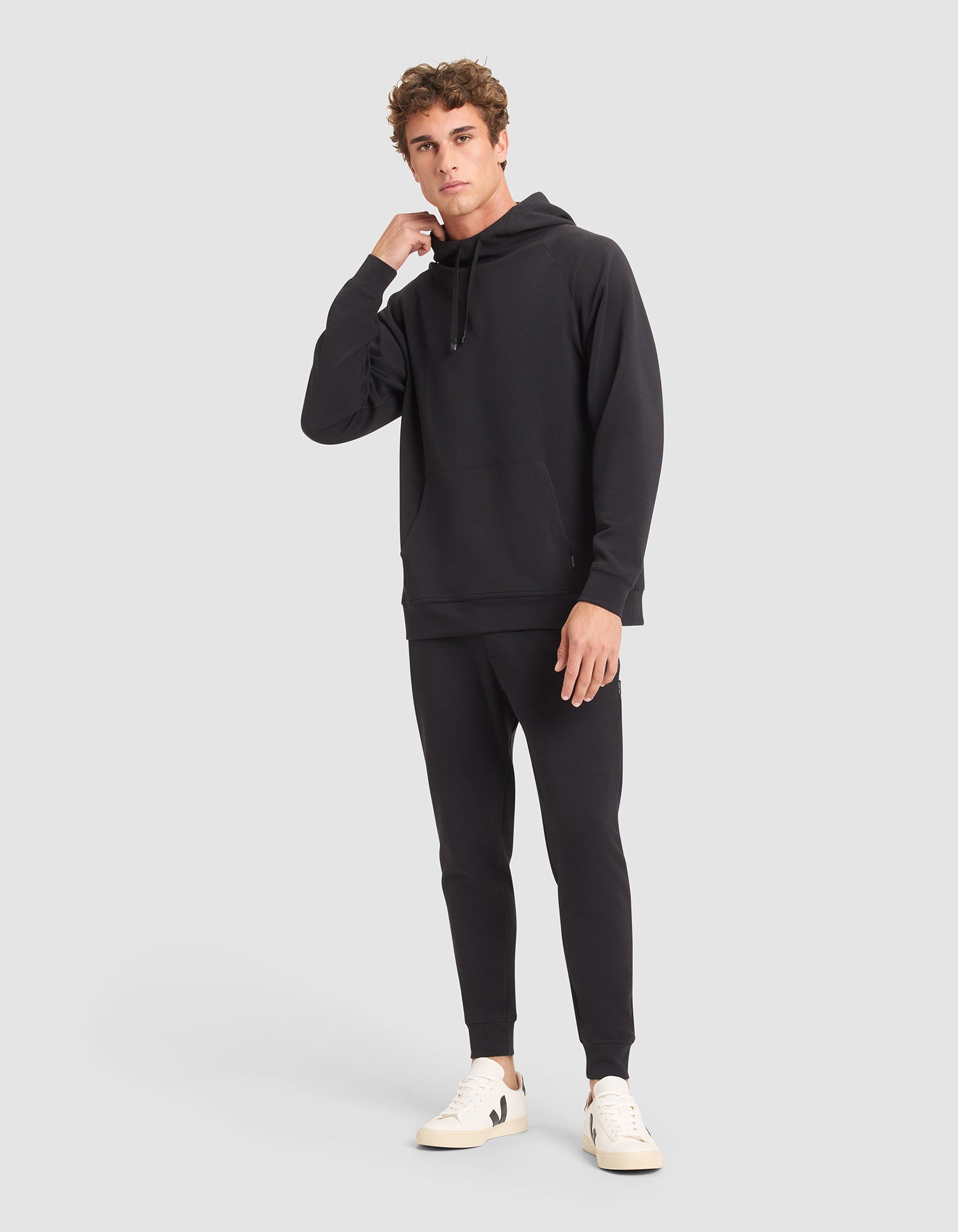 A young man with short curly hair poses against a light gray background, wearing the Cozy Earth Men's StretchTech Hoodie & Jogger Set in black with white sneakers, looking at the camera and touching his neck. 