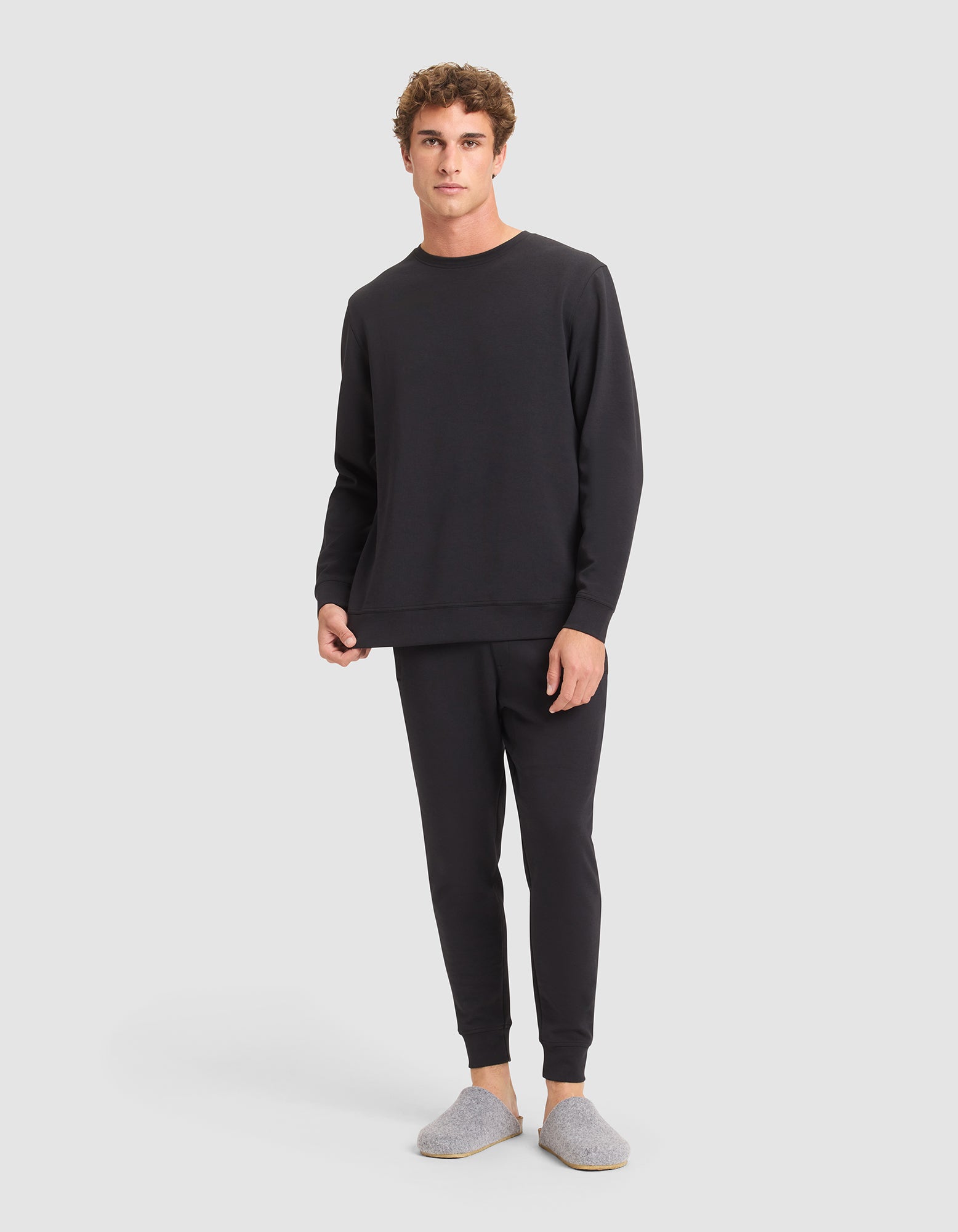 A man faces forward in a relaxed pose, wearing the Cozy Earth Men's StretchTech Crewneck & Jogger Set in black along with gray slippers, set against a plain background. 