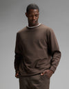 A man wearing the Cozy Earth Men’s StretchTech Crewneck in Cacao, paired with matching pants, stands with one hand in his pocket and a neutral expression against a plain gray background.|Color:Cacao