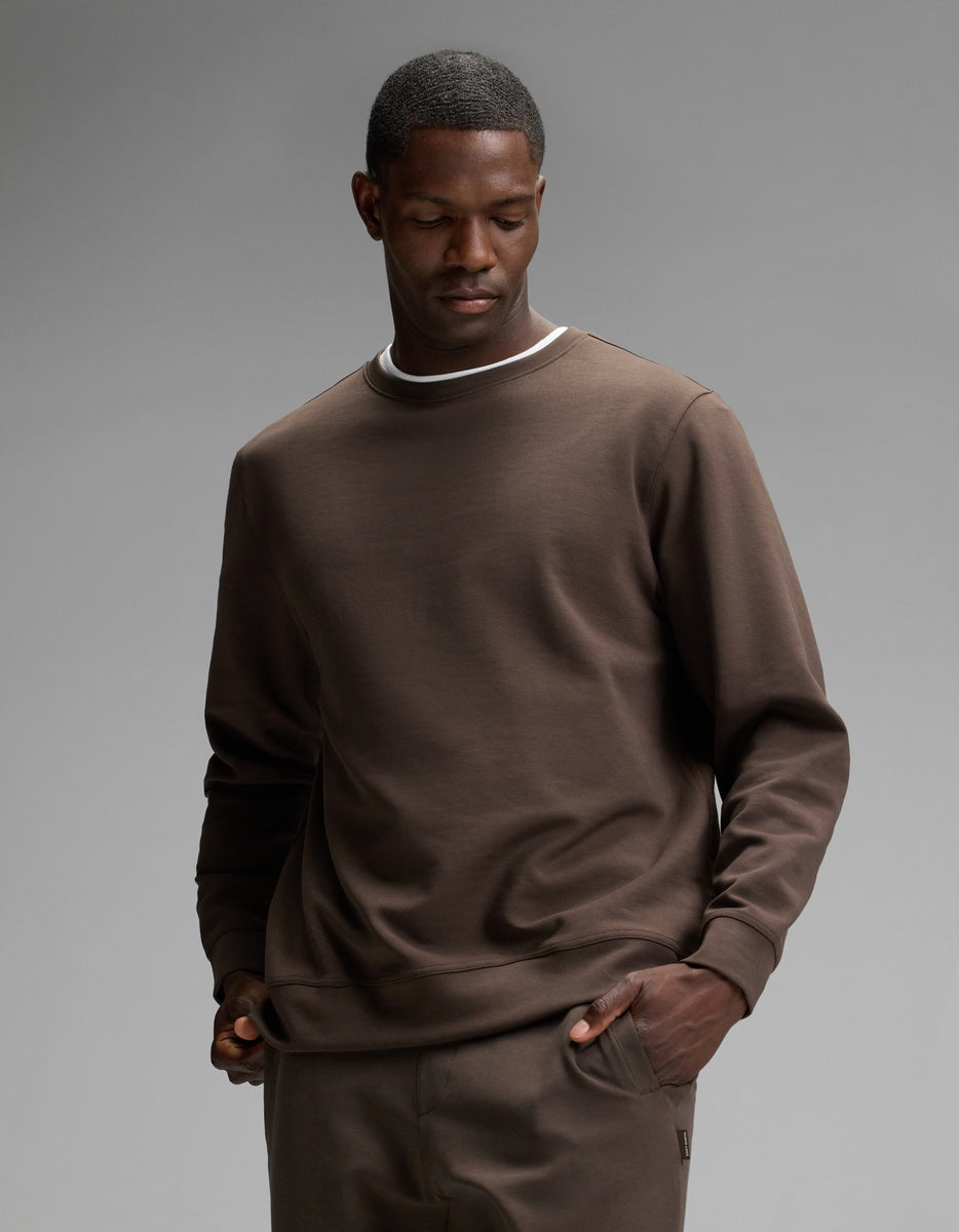 A man wearing the Cozy Earth Men’s StretchTech Crewneck in Cacao, paired with matching pants, stands with one hand in his pocket and a neutral expression against a plain gray background.