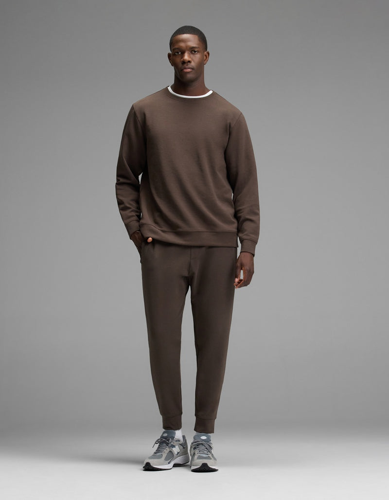 A man stands facing forward, wearing the Cozy Earth Men’s StretchTech Crewneck sweatshirt and matching jogger pants with gray sneakers, against a plain gray background.