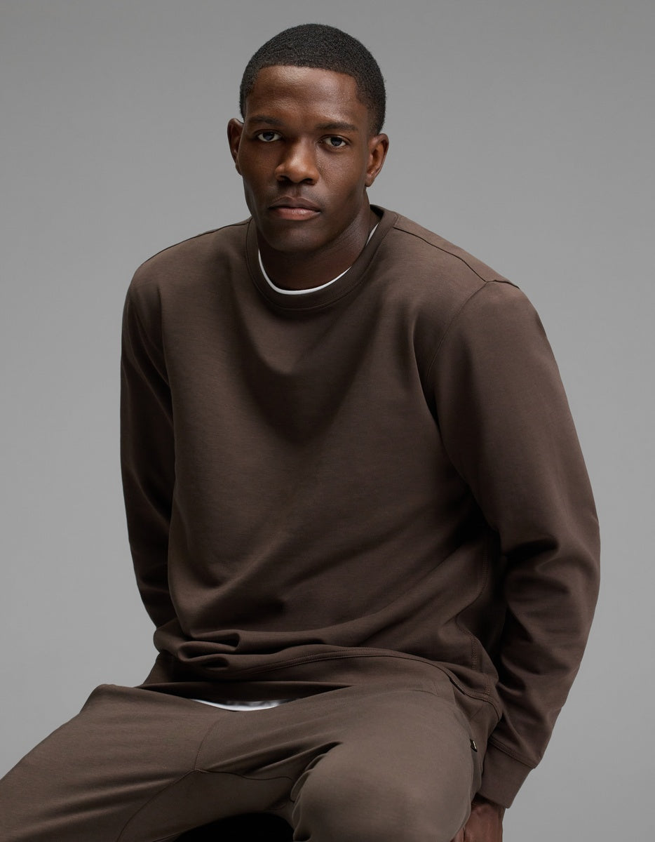 A man sits on a stool wearing the Cozy Earth Men’s StretchTech Crewneck and matching pants, looking at the camera with a neutral expression against a plain gray background.