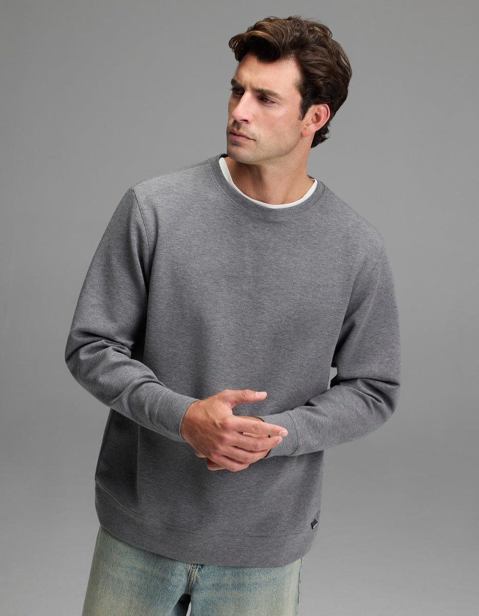 A man with short brown hair wears the Cozy Earth Men’s StretchTech Crewneck in Coal Heather and light blue jeans, standing against a plain gray background while looking to the side with his hands loosely clasped.