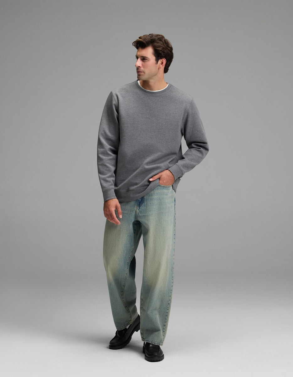 A man wearing the Cozy Earth Men’s StretchTech Crewneck and loose-fitting blue jeans stands against a plain gray background, looking to his left with one hand in his pocket.