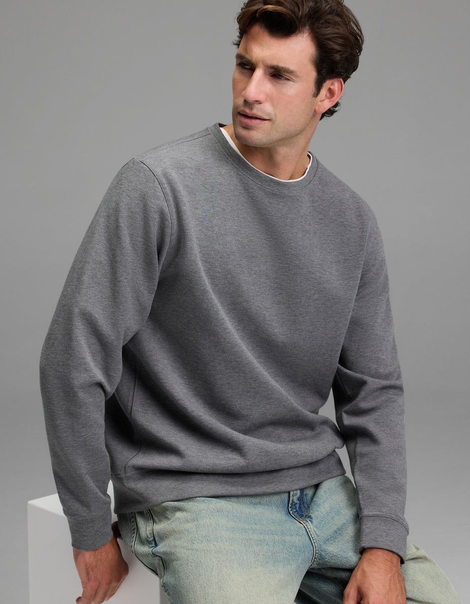 A man with brown hair wears the Cozy Earth Men’s StretchTech Crewneck in gray and light blue jeans while sitting on a white block against a plain gray background.