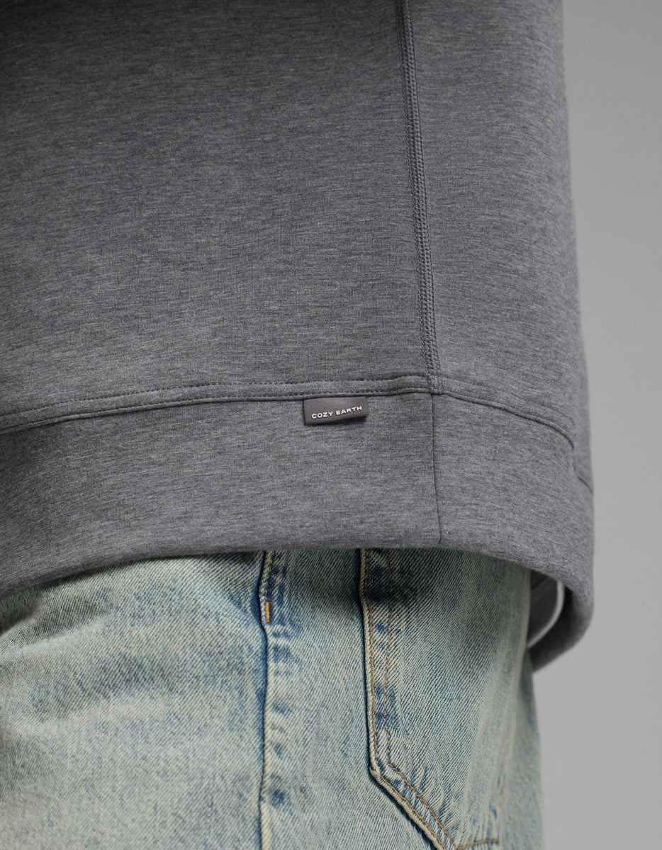 Close-up of a person wearing the Cozy Earth Men’s StretchTech Crewneck in gray and light blue jeans. The crewneck features a small rectangular tag near the hem with the text “COZY EARTH.”.