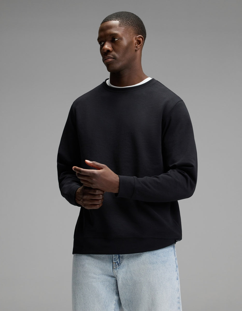 A person wearing the Cozy Earth Men’s StretchTech Crewneck in Jet Black over a white shirt and light blue jeans stands against a plain gray background, looking slightly to the side with hands together.